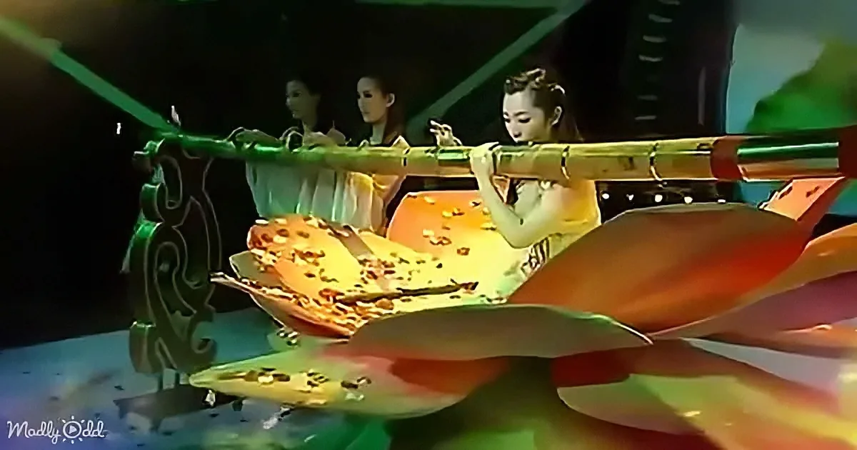 A performer in a flowing costume is holding a long, ornate object, possibly a musical instrument, on a stage with vibrant...