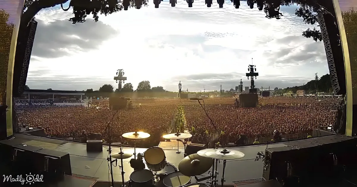 A massive crowd fills a concert stage under a cloudy sky, with a drum set in the foreground.