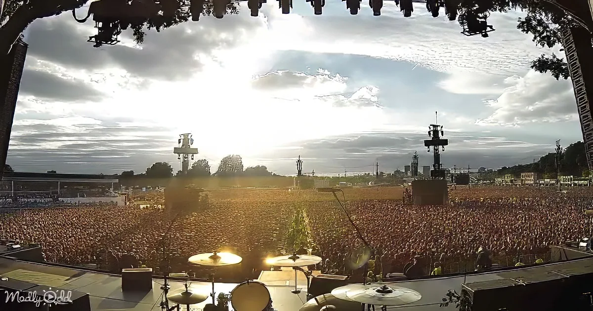 A massive crowd fills a festival stage under a dramatic sky, with sunlight breaking through.