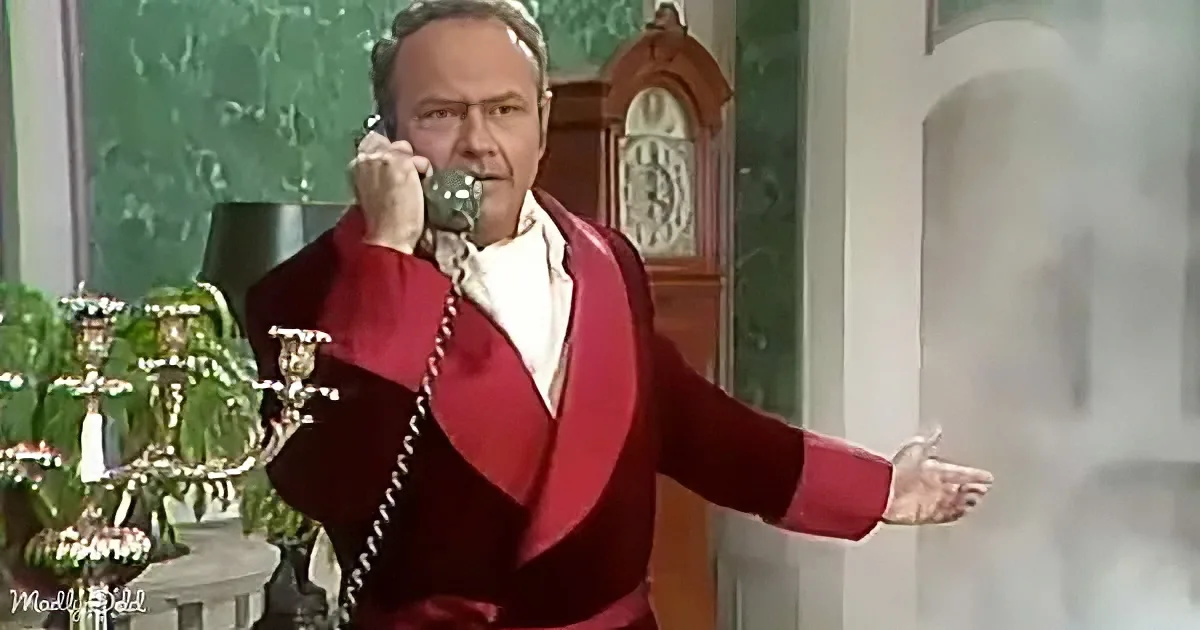 A man in a red robe talks on a rotary phone, gesturing with his other hand.