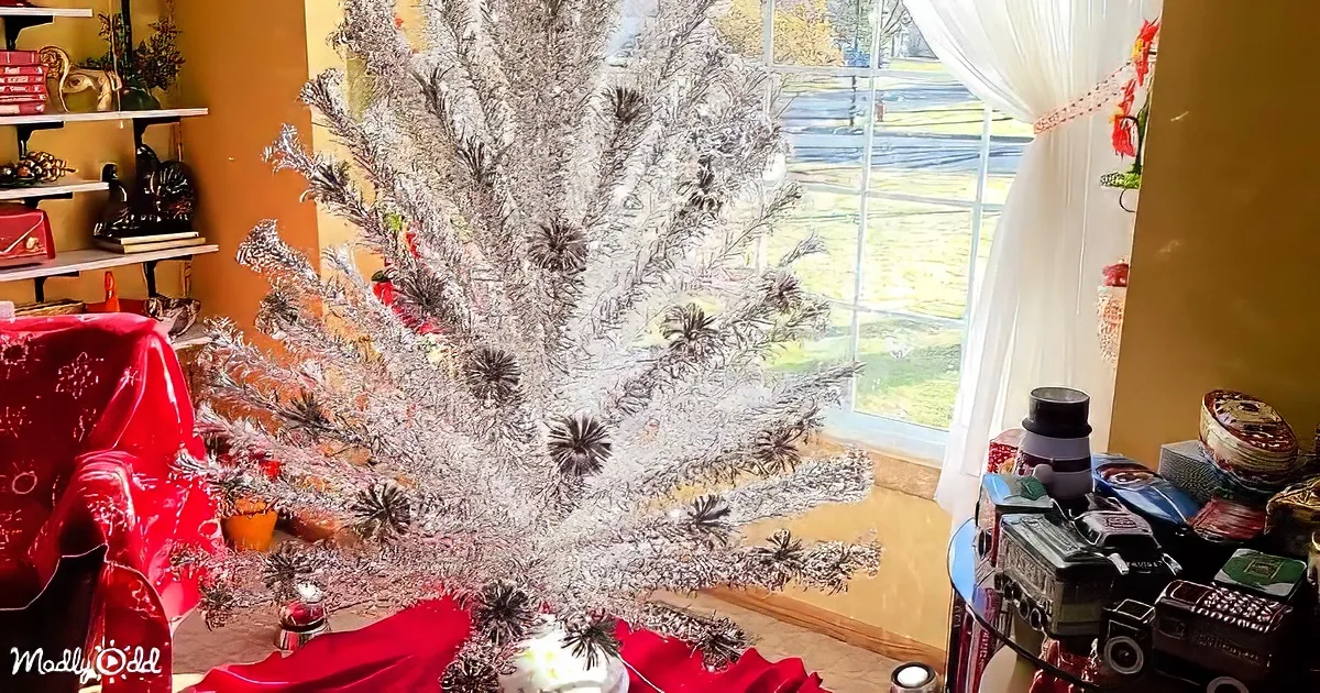 A silver Christmas tree adorned with ornaments stands in a room with a red tablecloth and a window view of a grassy yard.