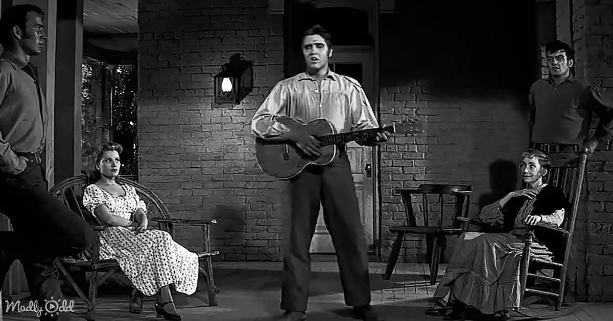 Elvis Presley plays guitar on stage, surrounded by people in a rustic setting.