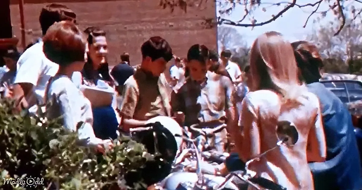 A group of people, including a man in a brown shirt, are gathered around a motorcycle in an outdoor setting.