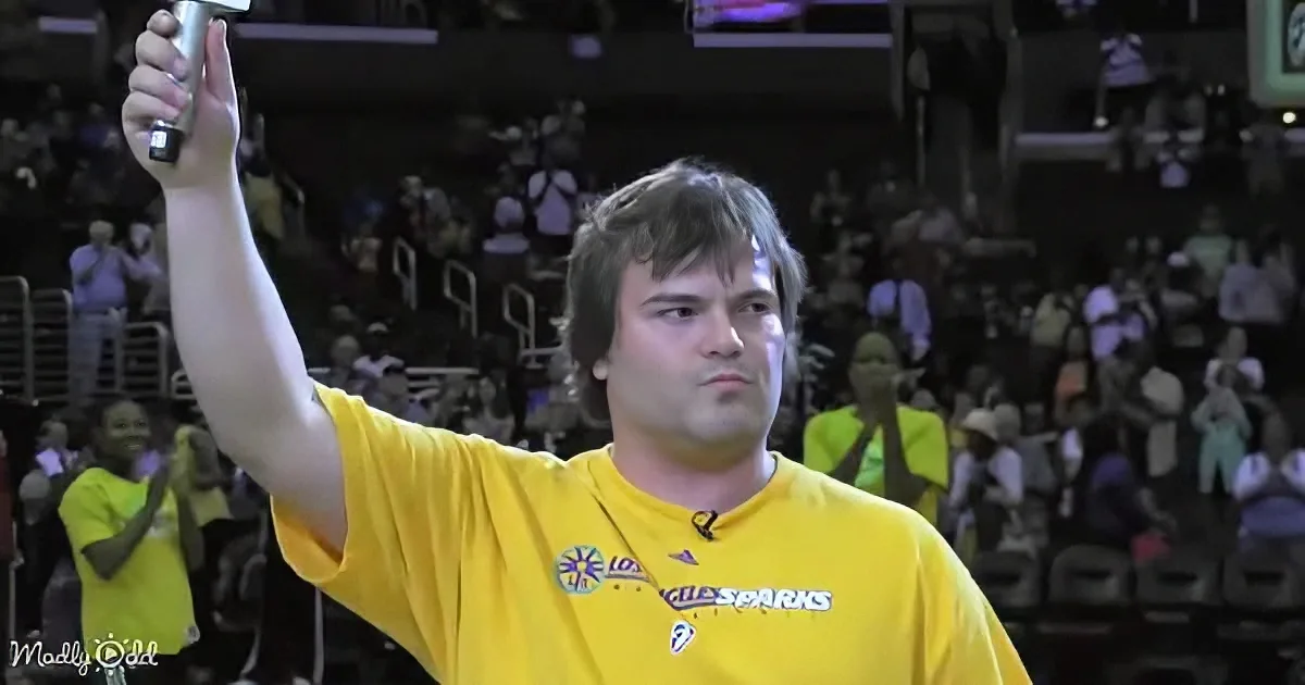 A man in a yellow Los Angeles Sparks shirt raises a microphone, likely addressing a crowd at a basketball game.