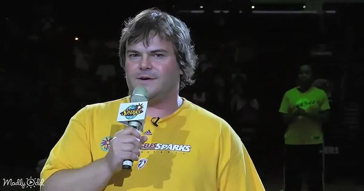 A man in a yellow "Los Angeles Sparks" shirt holds a microphone, speaking on stage.