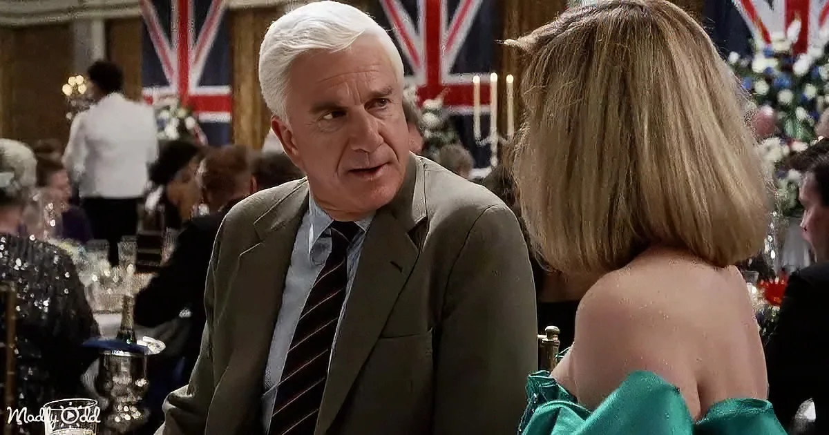 A man in a suit and tie is engaged in conversation with a woman in a formal setting with Union Jack flags in the background.