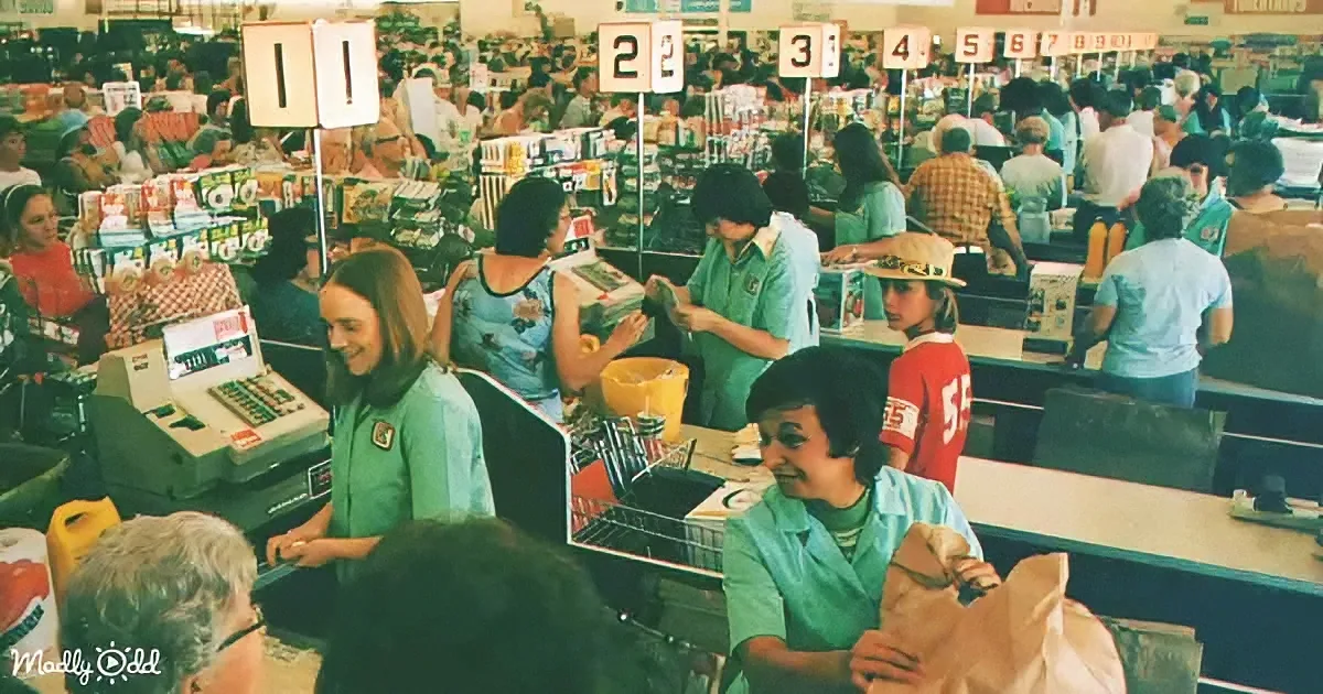 Cashiers in green shirts smile as customers wait in line at a busy grocery store checkout.