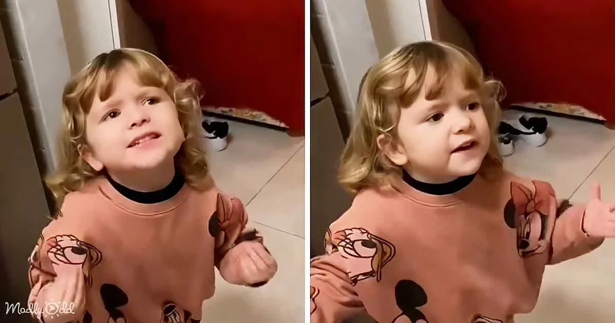 A toddler in a pink Mickey Mouse shirt stands indoors, arms outstretched, with a curious expression.