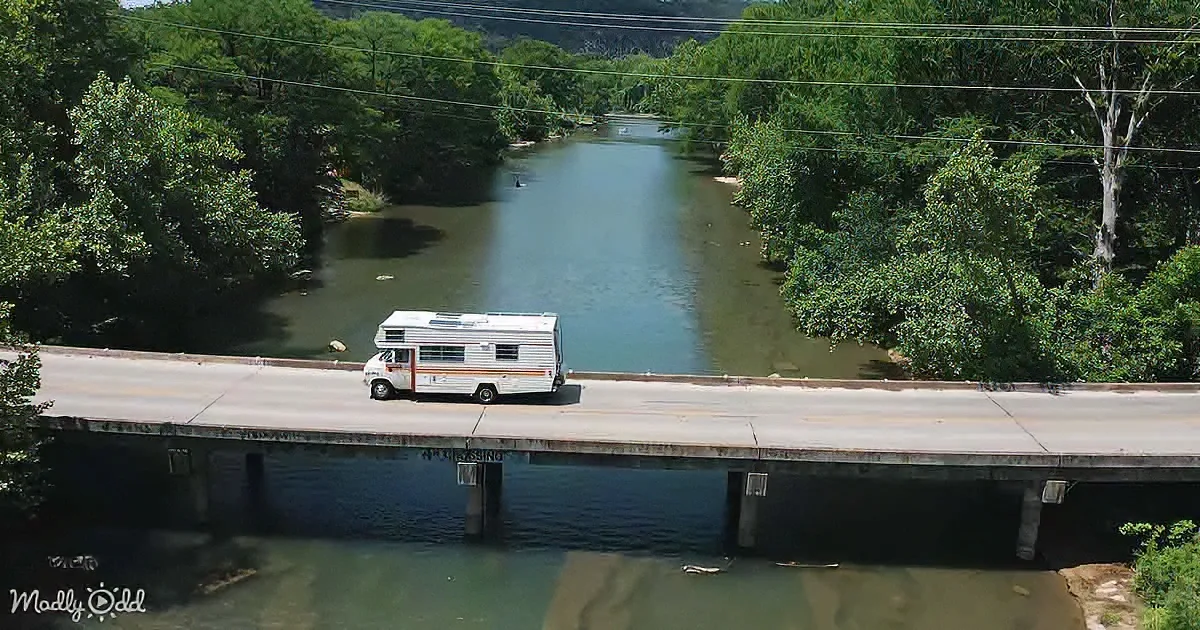A white RV with orange stripes drives across a bridge over a calm river surrounded by lush green trees.