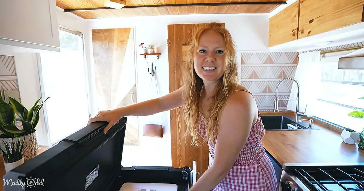 A woman in a red checkered dress smiles in a cozy kitchen with wooden cabinets and a black stove.