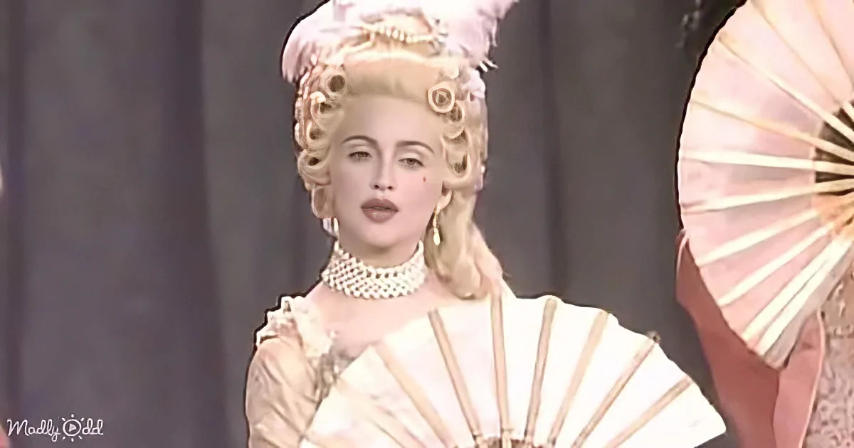 A person in a historical costume with elaborate hair and jewelry, holding a fan.
