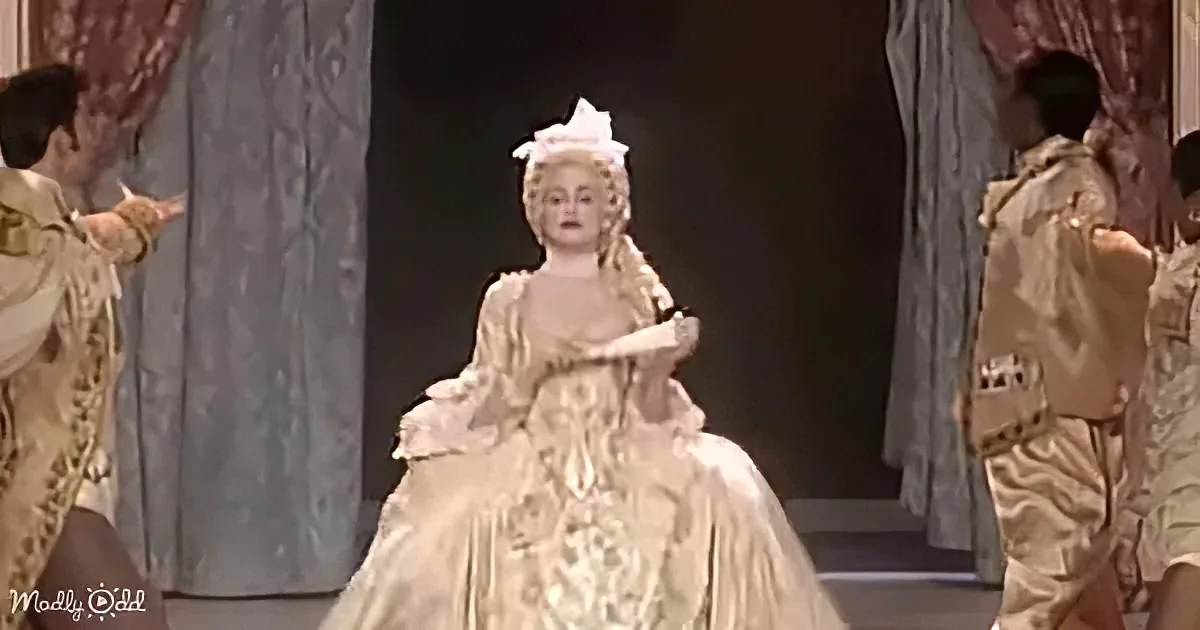 A woman in an elaborate, cream-colored, historical gown with a large, ornate headdress stands center stage.