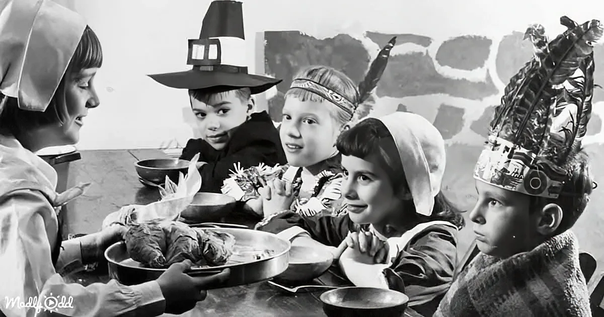 Children in Pilgrim and Native American costumes gather around a table with food, celebrating Thanksgiving.