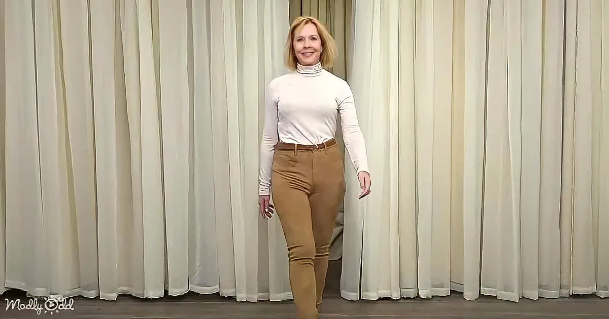 A woman in a white turtleneck and brown pants stands in front of a cream-colored curtain backdrop.