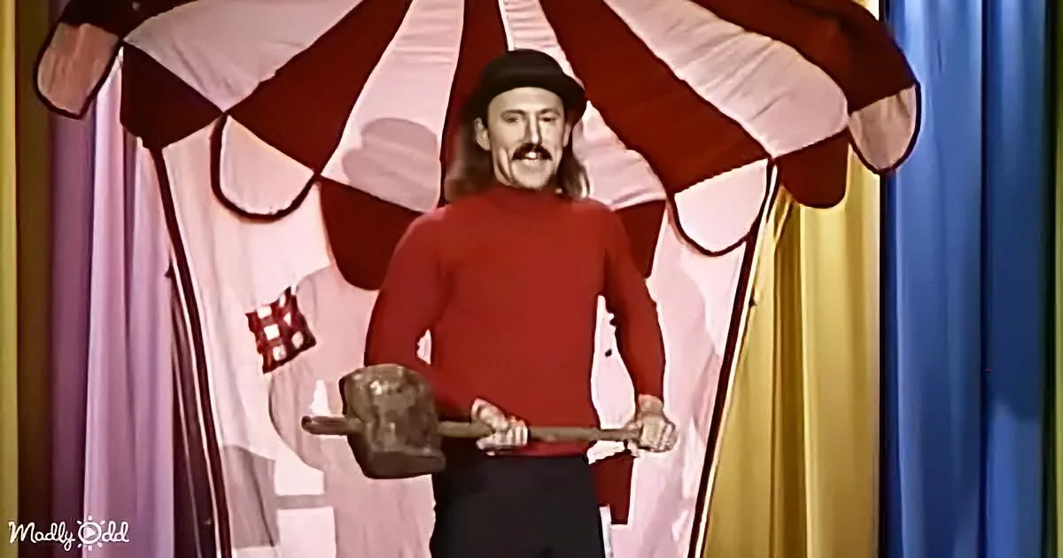 A man in a red sweater and bowler hat holds two hammers, standing in front of a colorful circus tent backdrop.