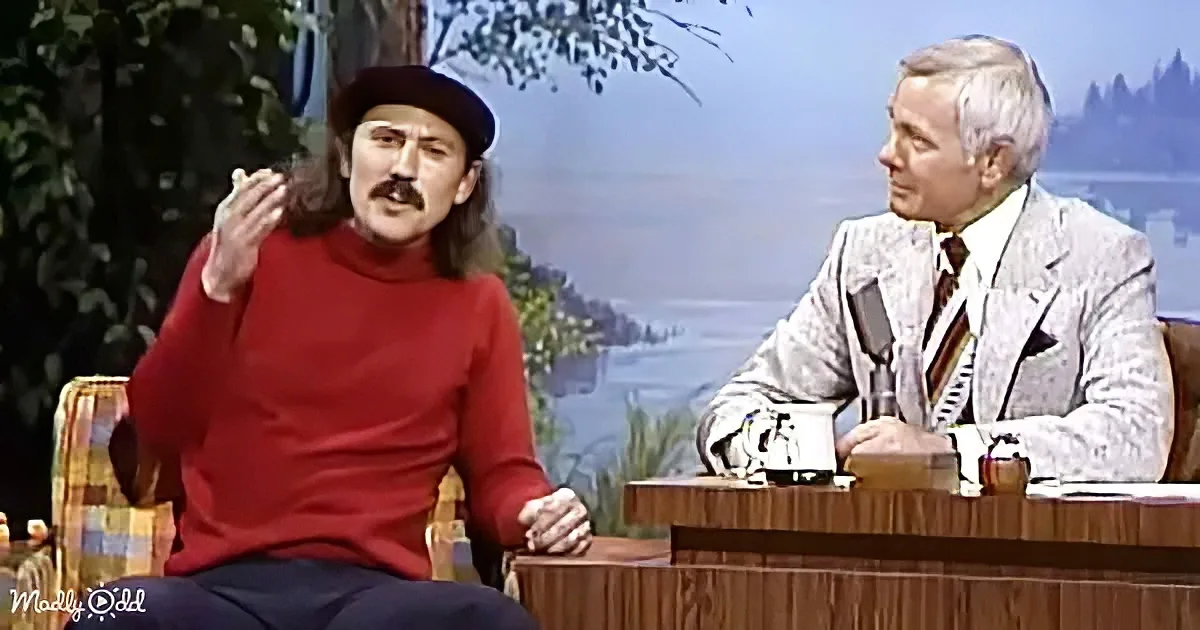 A man in a red sweater and hat gestures while seated, talking to a man in a suit on a talk show set.