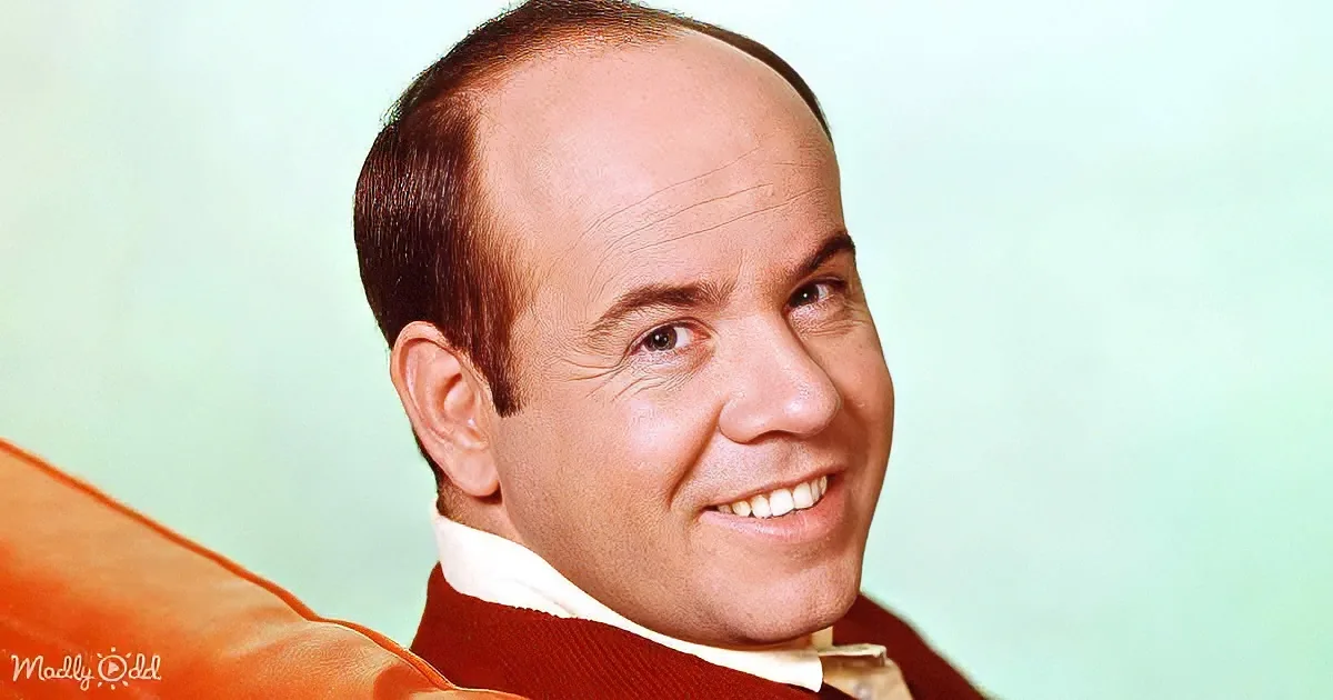 A man with a balding head, wearing a red sweater and white shirt, smiles warmly against a light green background.