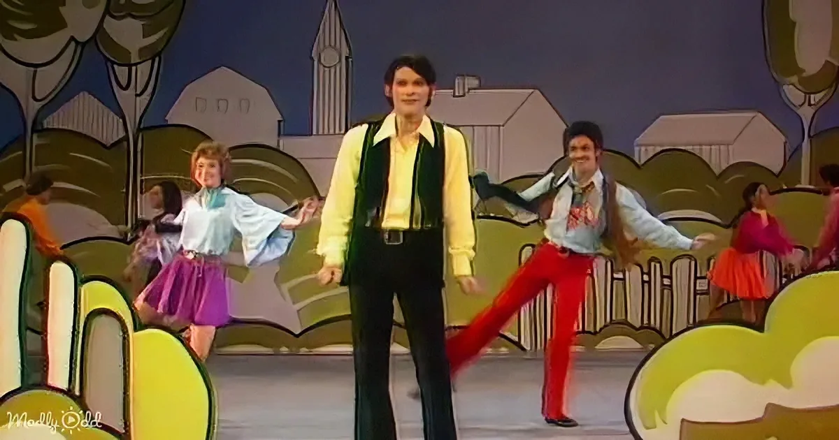 A man in a green vest and yellow shirt dances on stage with a colorful backdrop.