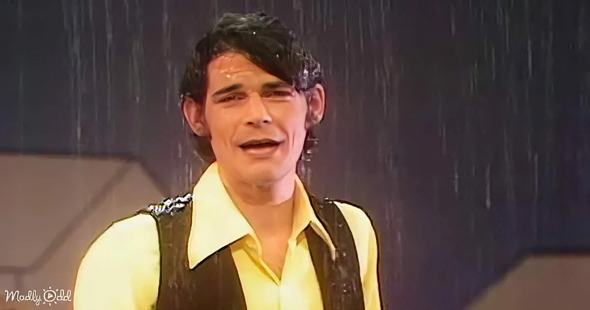 A man in a yellow shirt and brown vest stands in the rain, looking up with a surprised expression.