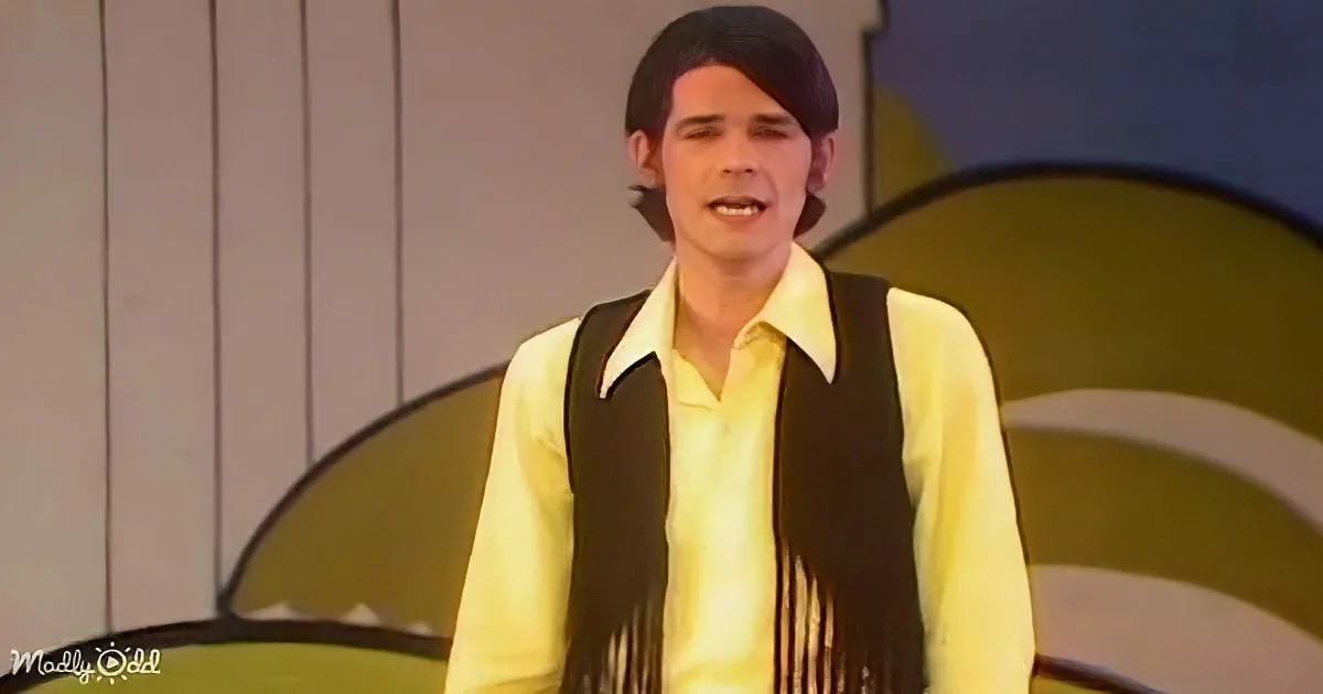 A person in a yellow shirt and black vest with fringes stands in front of a green and white background.