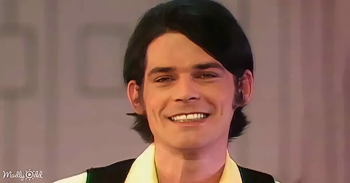 A smiling man with dark hair, wearing a white and black outfit, against a blurred background.