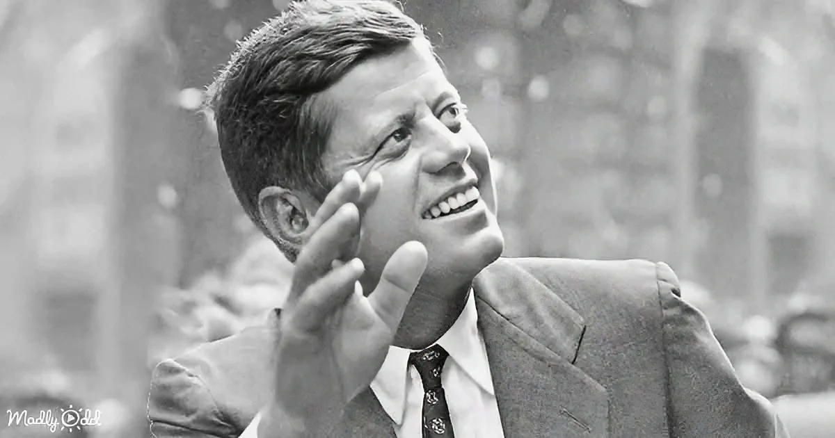 A man in a suit and tie gestures with his right hand, smiling, in a black-and-white photo.