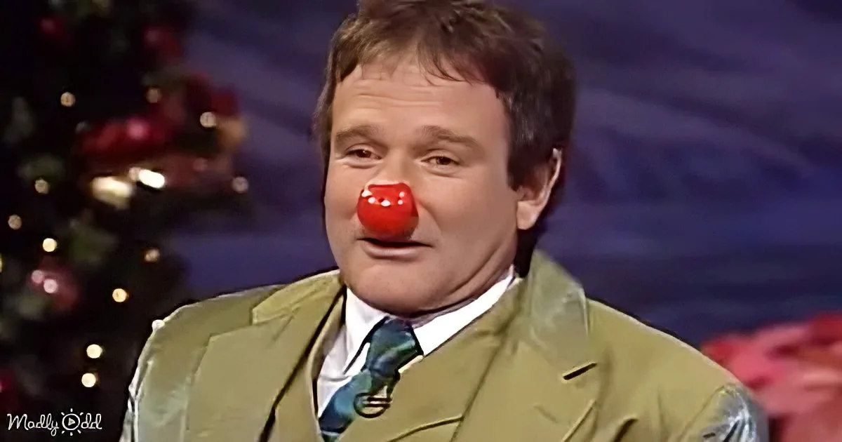 A man in a green suit with a red clown nose, sitting in front of a Christmas tree.