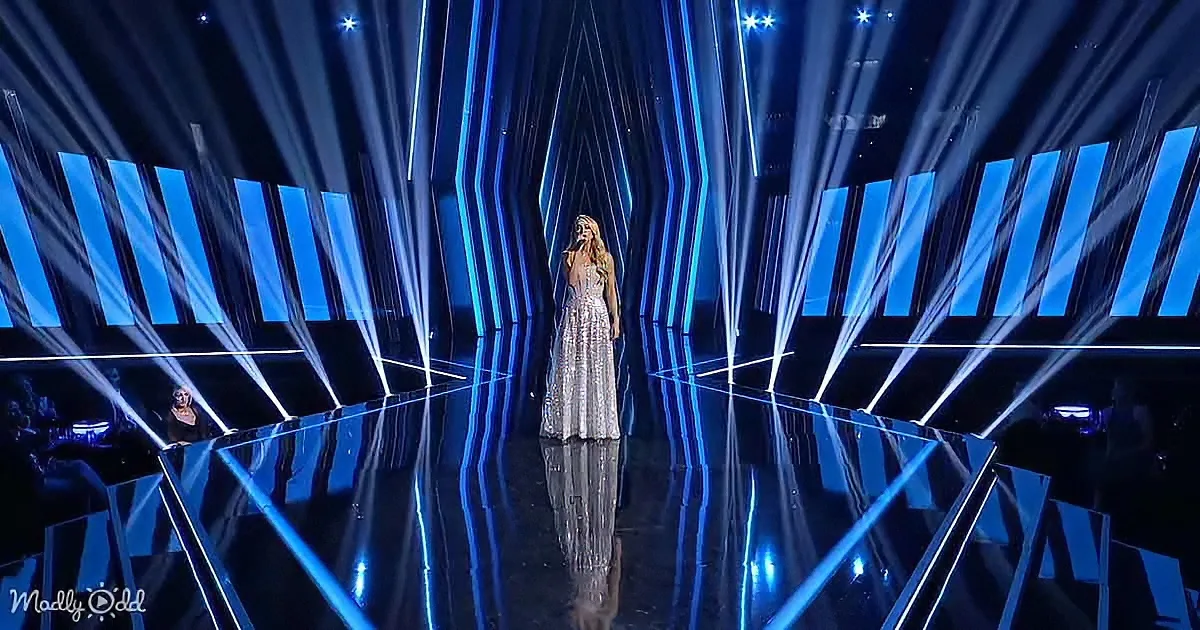 A woman in a silver dress sings on a stage with blue lighting and geometric patterns.