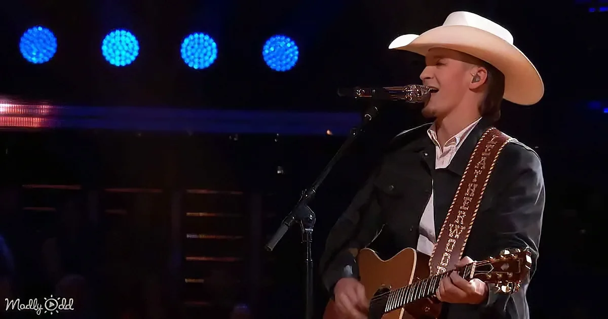 A man in a cowboy hat and black jacket plays a guitar on stage with blue lights in the background.