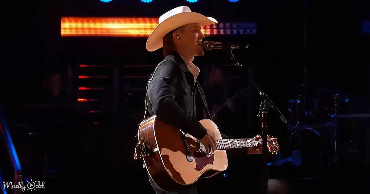 A person in a white cowboy hat plays an acoustic guitar on stage with a microphone.