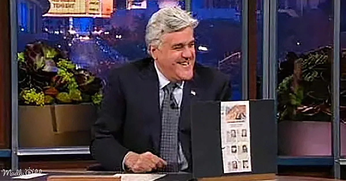 A man in a suit sits at a desk, smiling, with a cityscape and plants in the background.