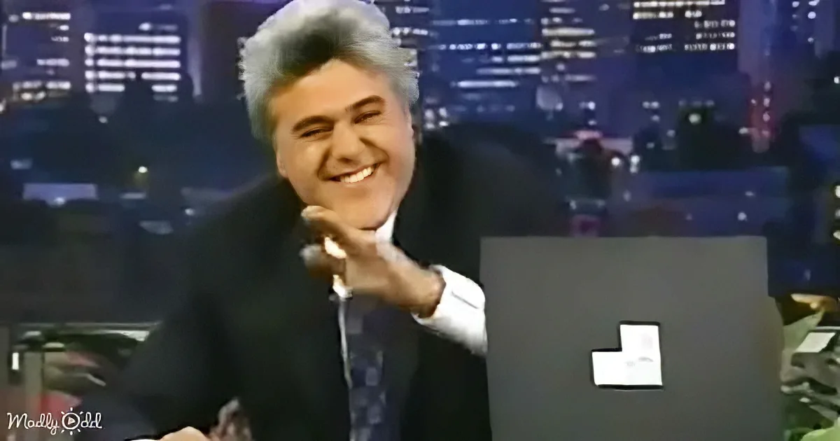 A man in a suit smiles while holding a microphone, seated at a desk with a cityscape backdrop.