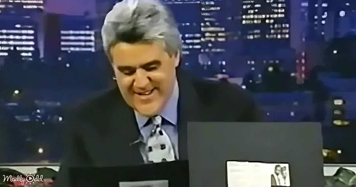 A man in a suit and tie is smiling while sitting at a desk with a cityscape background.