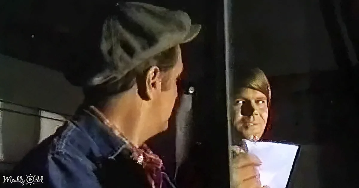 A man in a cowboy hat and denim jacket stands facing another person in a dark room.