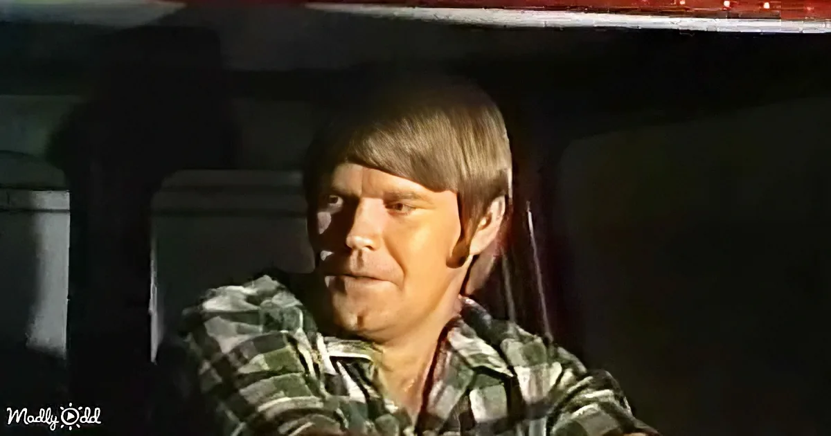 A man in a plaid shirt sits inside a vehicle, looking to the side with a serious expression.