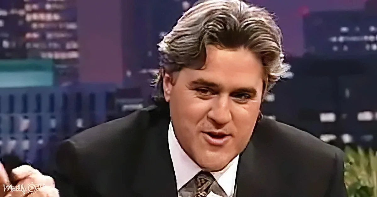 A man in a suit and tie is speaking, holding a microphone, with a cityscape background.