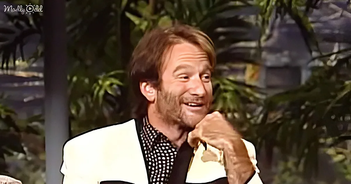 A man in a white suit jacket and checkered shirt is smiling and gesturing with his hand.