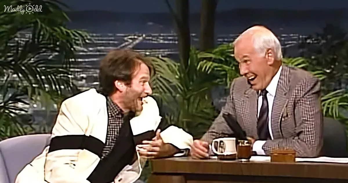 Two men, one in a white suit, are laughing on a talk show set with a cityscape backdrop.