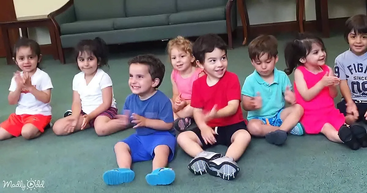 Children in colorful clothes sit on the floor, clapping and smiling.