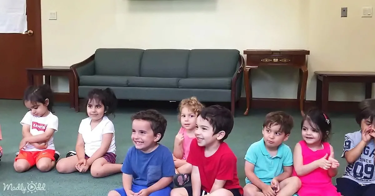 Children sit on the floor in a row, smiling and looking at something off-camera.