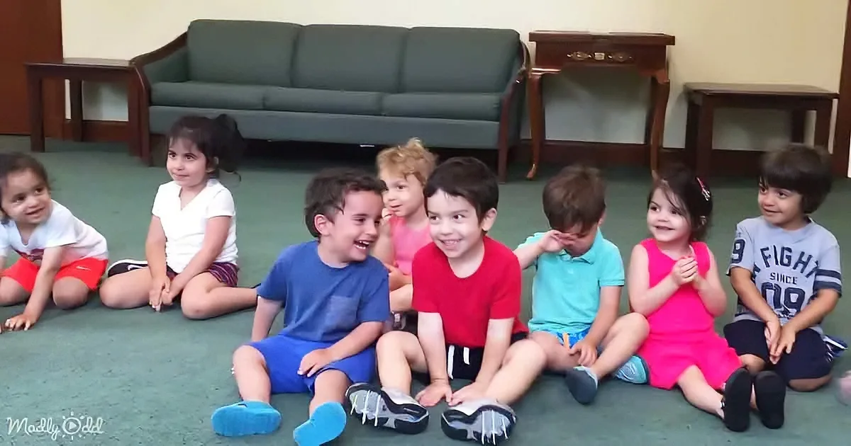 Children sit in a row on the floor, smiling and looking at the camera.