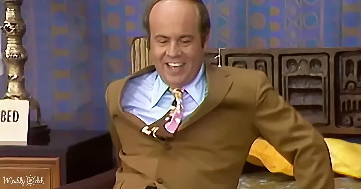A man in a brown suit and tie is sitting in a chair, smiling widely, with a "BED" sign visible in the background.