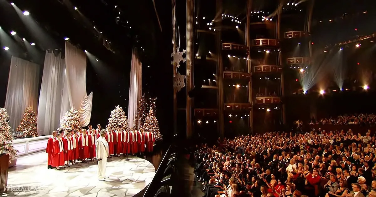 A choir in red and white stands on stage with Christmas trees, surrounded by a large audience in a theater.