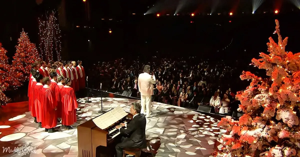 A man in a white suit leads a choir in red robes on a stage with Christmas trees and a large audience.