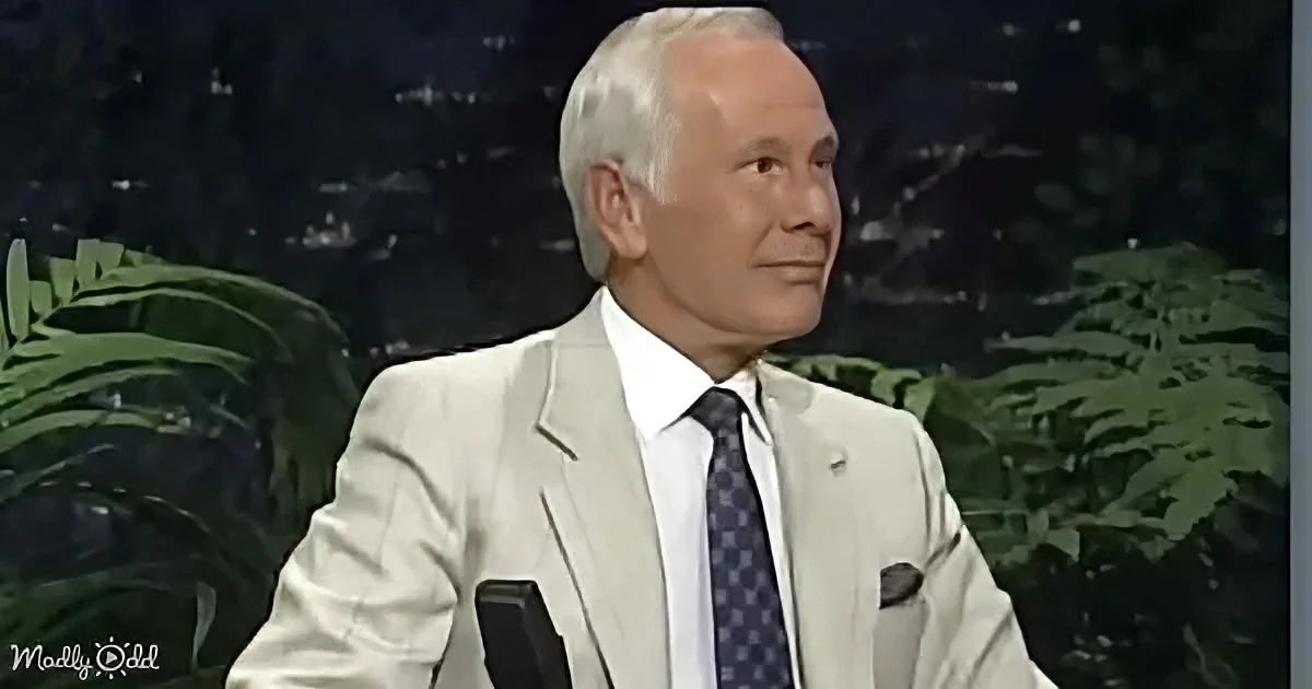 A man in a light suit and tie sits on a talk show set with a cityscape backdrop.