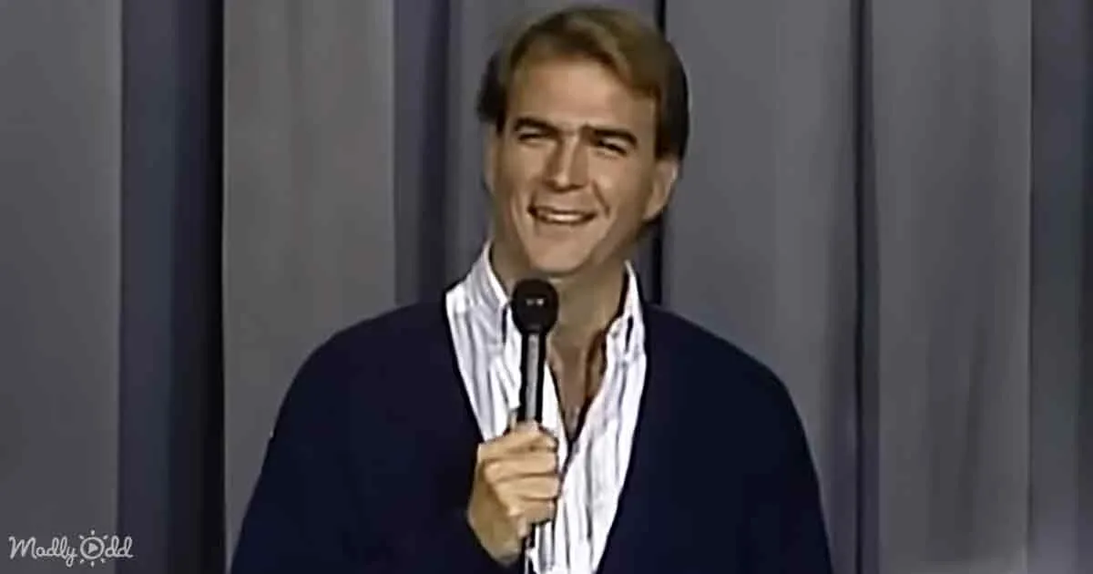 A man in a striped shirt and navy sweater holds a microphone, smiling on stage.