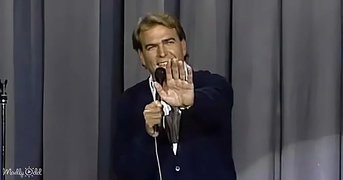A man in a suit holds a microphone and gestures with his hand, appearing to speak or perform.