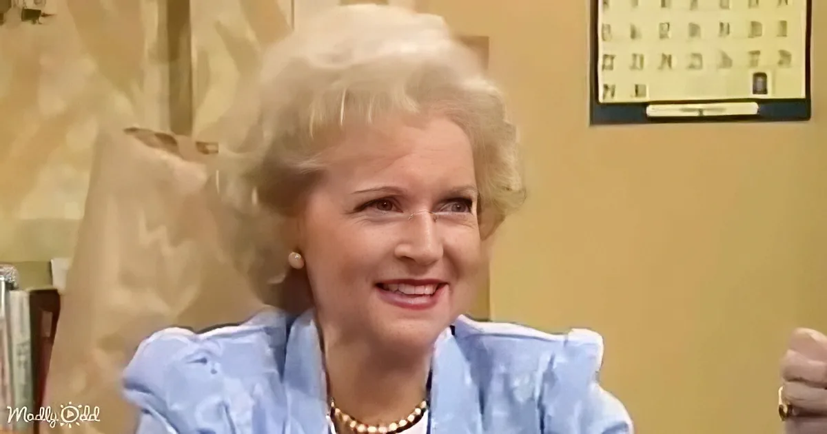 A smiling woman with blonde hair, wearing a blue dress and pearl necklace, sits in a room with a calendar on the wall.