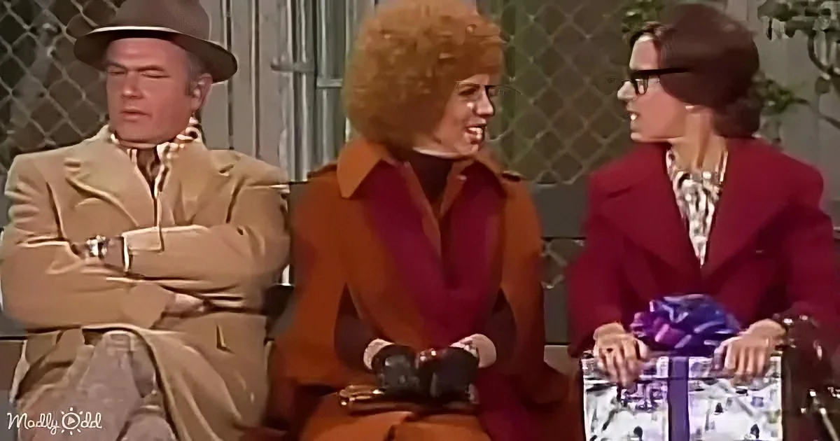 A woman with an afro hairstyle in a brown coat sits between two other women, one holding a gift, in a vintage setting.