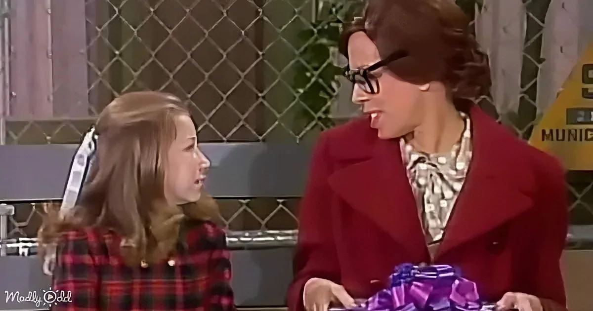 A woman in a red coat and glasses smiles at a girl in a plaid shirt, holding a wrapped gift.